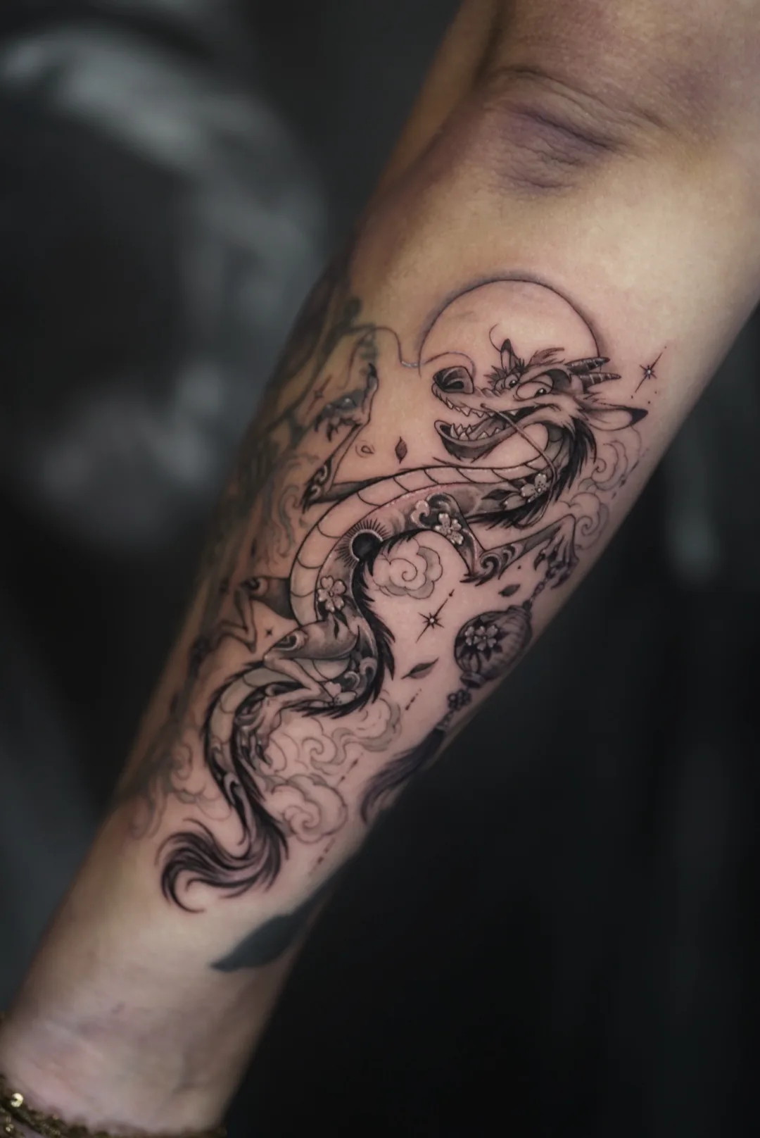 A fine-line dragon winding across a forearm, with a full moon, clouds, blossoms and a hanging lantern.