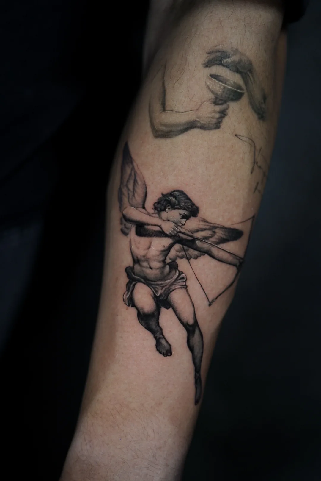 A micro-realism tattoo of a winged cupid drawing his bow, on a forearm.